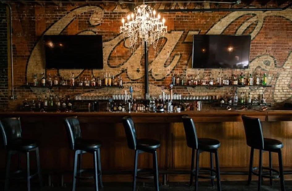 The bar under a crystal chandelier with the Coca-Cola ghost sign painted on the brick behind it